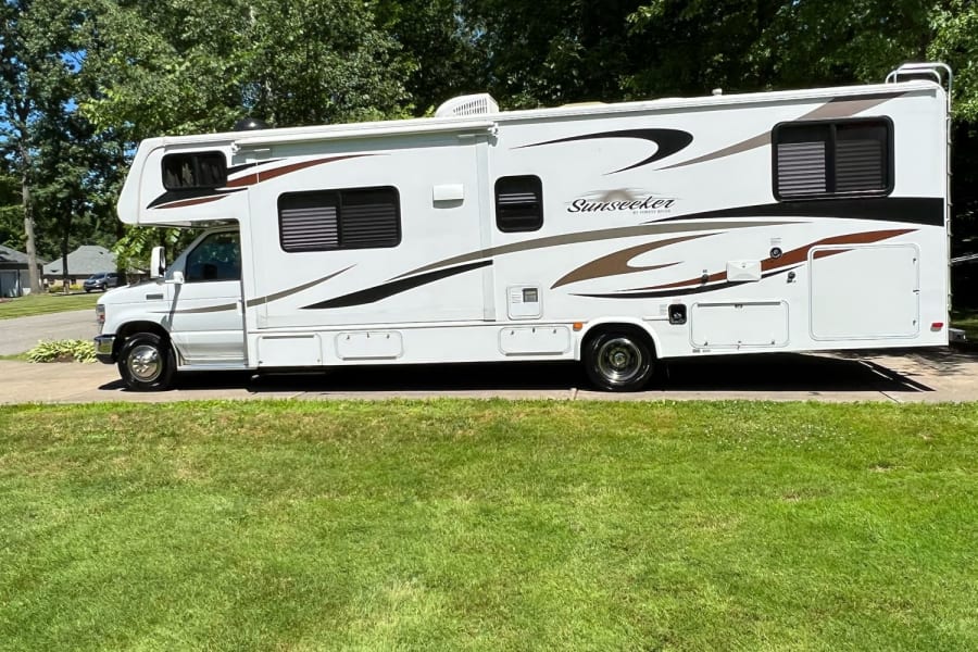 A white Sunseeker Class C RV, ideal for an rv rental, is parked on a driveway with green grass and trees in the background.