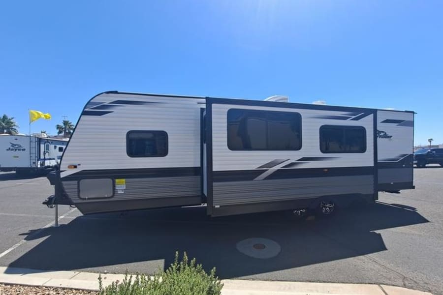 White & gray travel trailer, an RV rental, with a slide-out, parked on a sunny day in a paved lot.