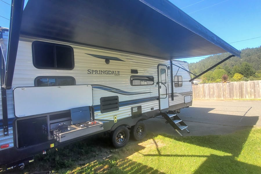 White Springdale RV rental with extended dark awning & pull-out outdoor kitchen, parked on grass.