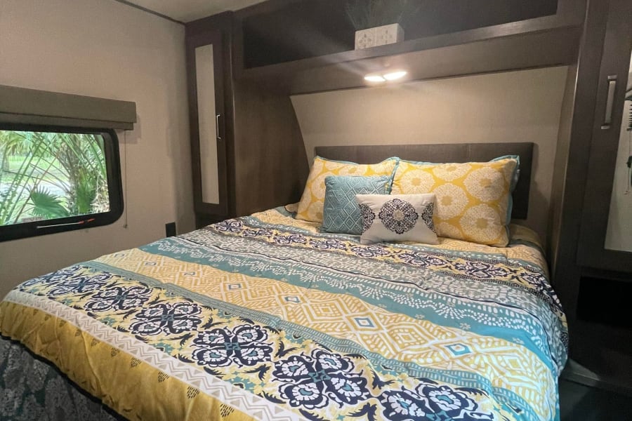 Cozy RV rental bedroom featuring colorful yellow/blue patterned bedding, throw pillows, and a bright window view.