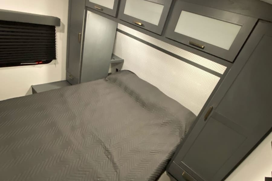 Modern RV rental bedroom with a grey bed, overhead cabinets, a patterned headboard, and built-in storage.