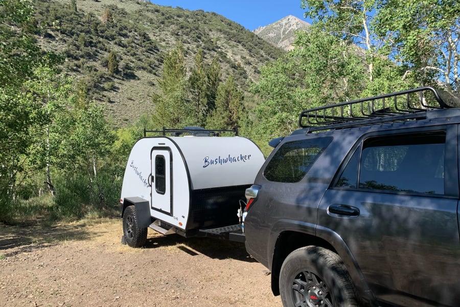 Explore nature with this RV rental: an SUV tows a teardrop trailer amidst green mountains. Perfect for your next camping trip.