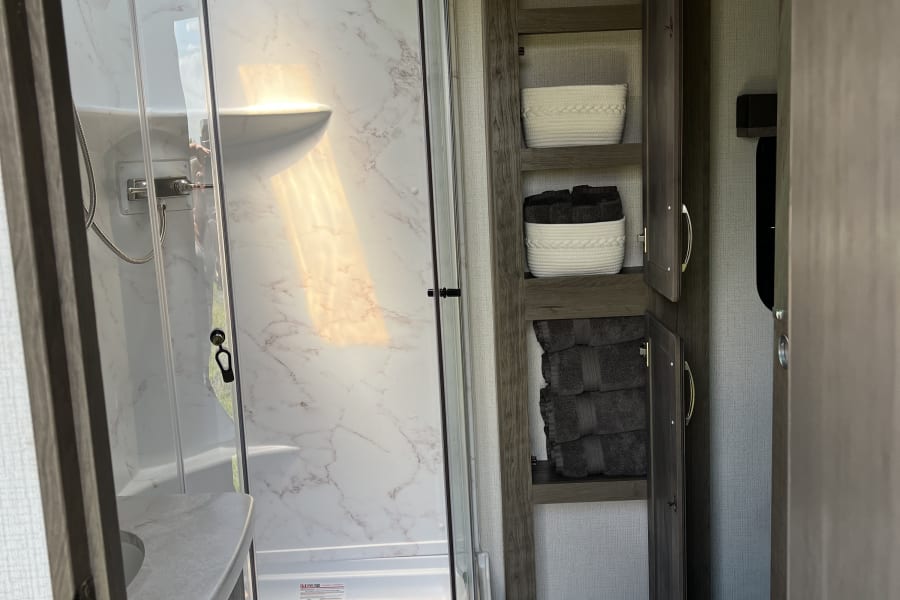 Bright RV rental bathroom with a modern corner shower, vanity, organized shelving for storage, and a toilet. Sunlight streams in.