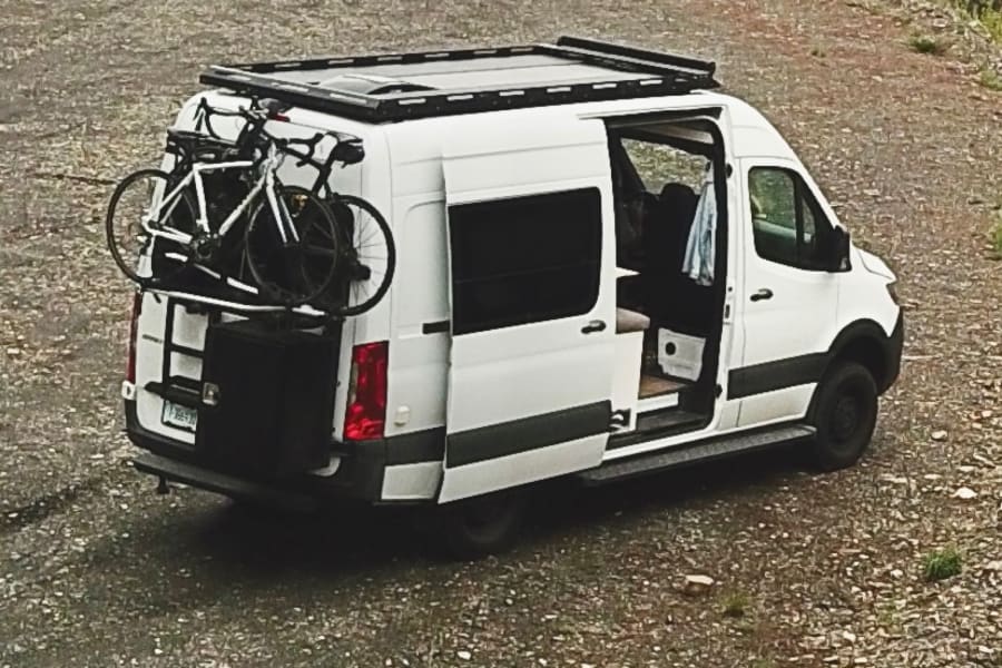 White adventure RV rental van, with bikes & open side door on gravel.