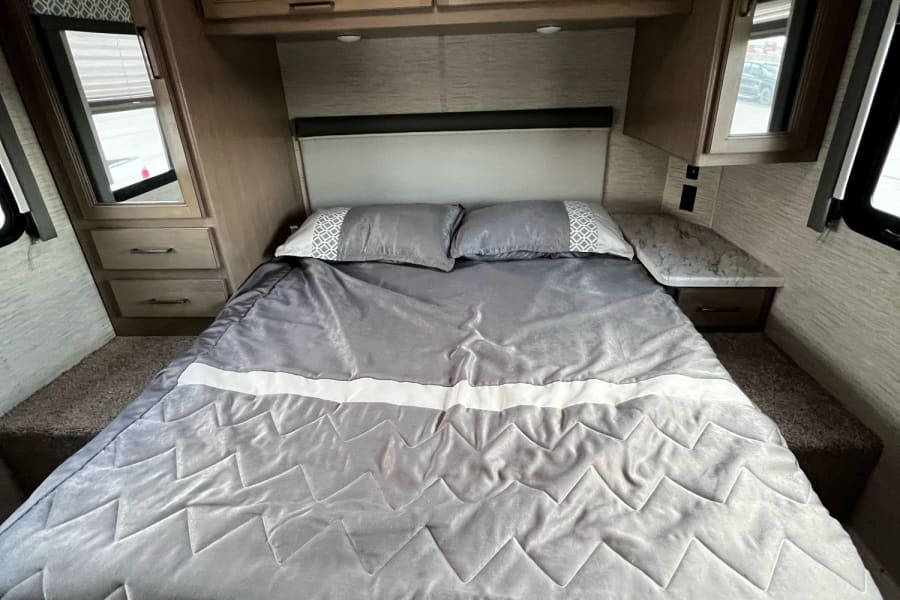 Cozy RV rental bedroom with a large gray bed, overhead cabinets, and side nightstands. Bright interior for your journey.