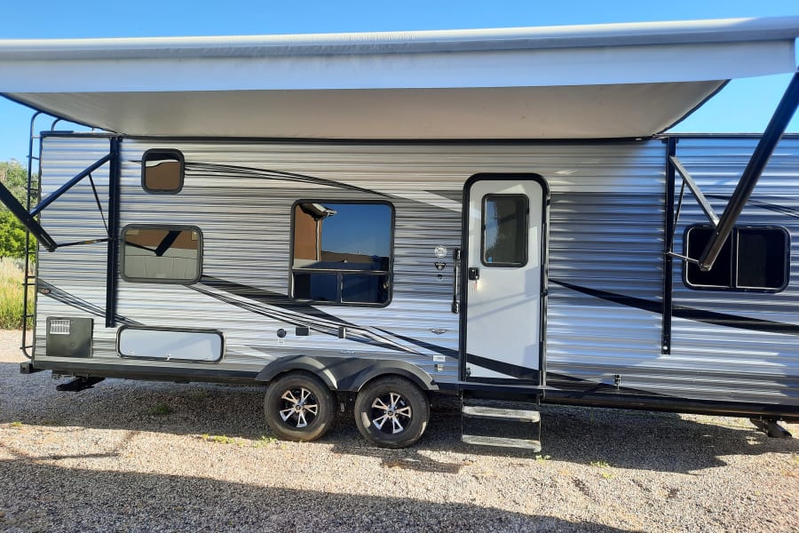Side view of a modern silver and grey travel trailer RV rental, featuring an extended awning, windows, door, and dual axles, ready for adventure.