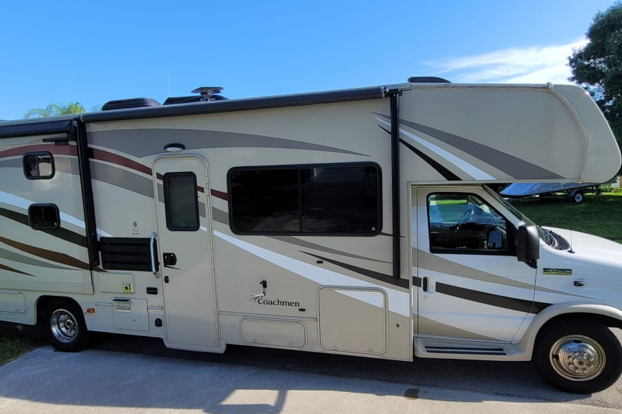 This Coachmen Leprechaun Class C RV could be your next RV rental. Shows the parked side of a beige RV with stripe graphics.