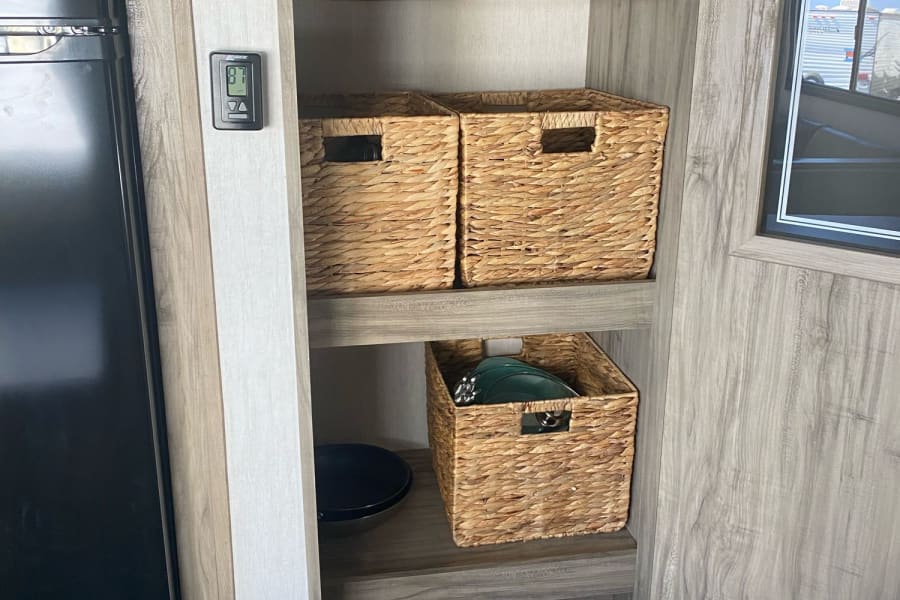 Inside an RV rental: a wood shelving unit with woven baskets, a thermostat, and a trash can for organized living.