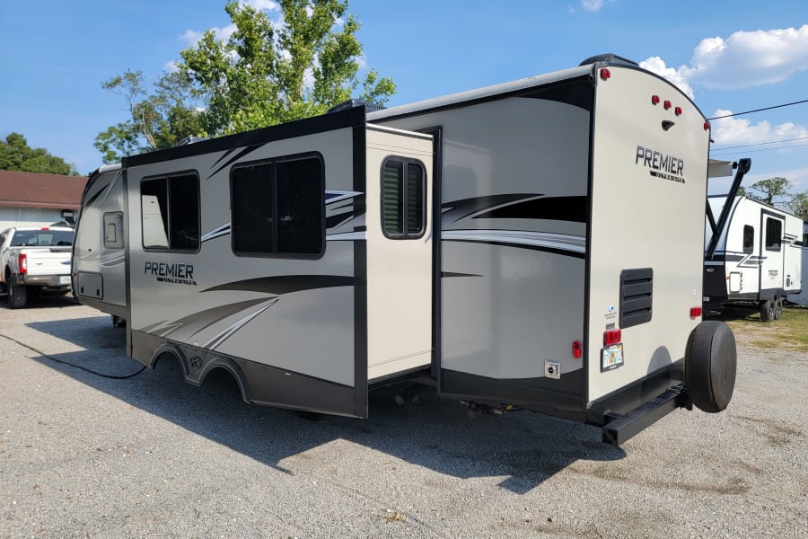 Light gray Premier Ultra Lite RV rental with slide-out extended, parked on a gravel lot under a blue sky.