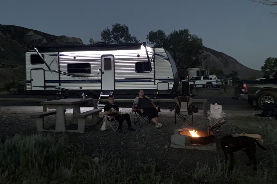 Relaxing RV rental night: Campers by a warm fire, with a large travel trailer and dog at dusk.
