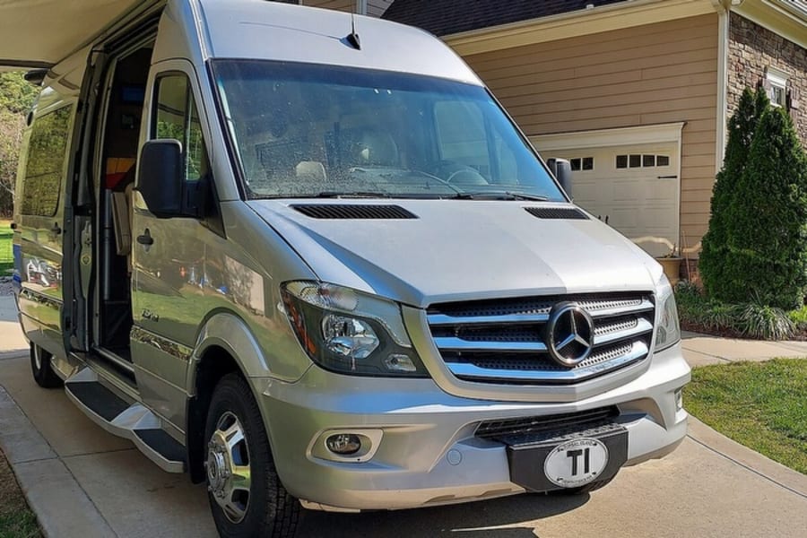 Silver Mercedes-Benz Sprinter RV rental parked on a suburban driveway with awning extended, showing interior.