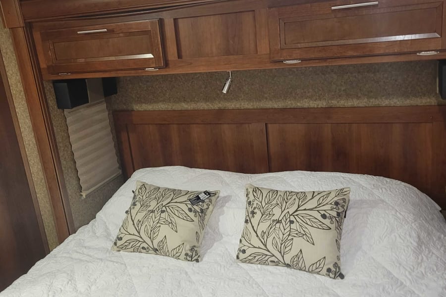 Relax in this cozy RV rental bedroom. Features a large bed with white quilted bedding, patterned pillows & wood overhead storage.
