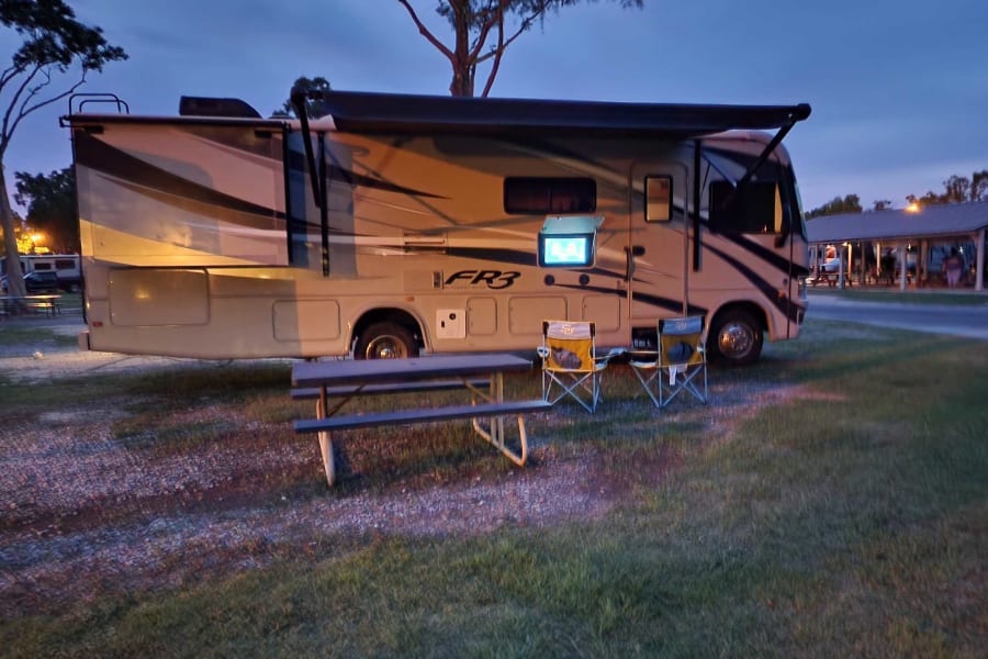 An RV rental provides comfort at dusk: a motorhome with an outdoor TV, chairs, and picnic table on a grassy site.