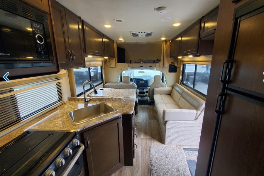 Bright RV rental interior showing a full kitchen with sink/stove, overhead cabinets, and a comfy sofa.