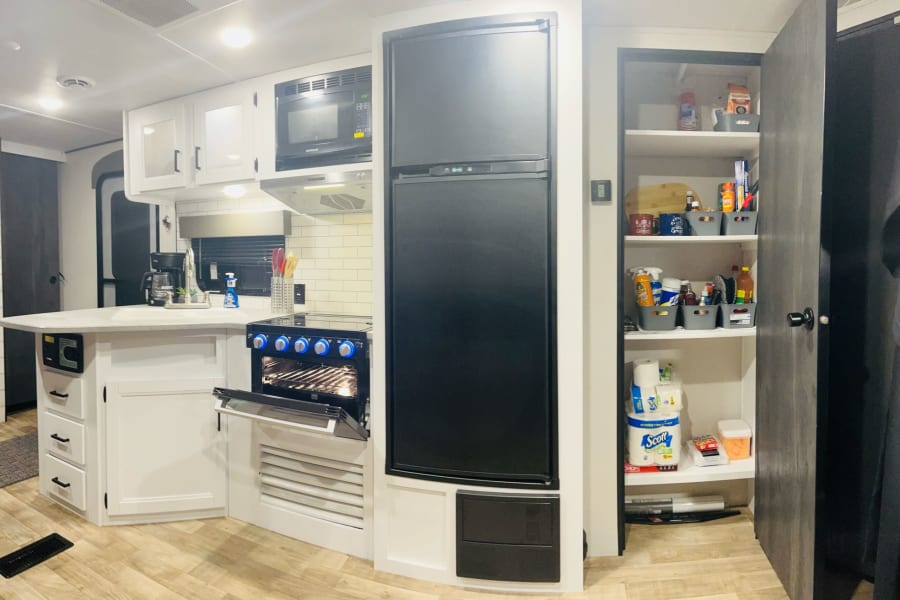 Modern RV rental kitchen with white cabinets, dark appliances, and an open, organized pantry on the right.