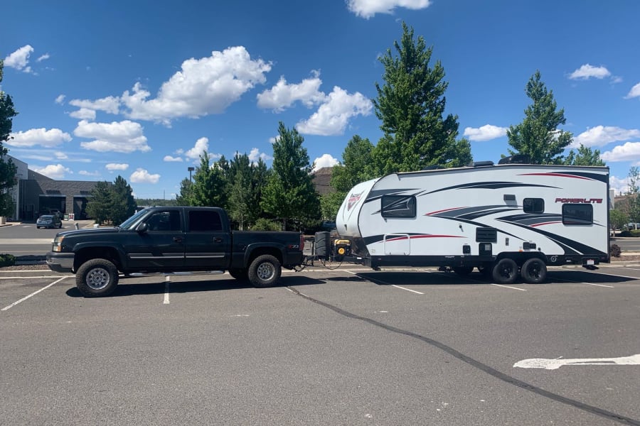 A dark pickup tows a white Powerlite travel trailer in a sunny parking lot. Ready for your rv rental road trip!