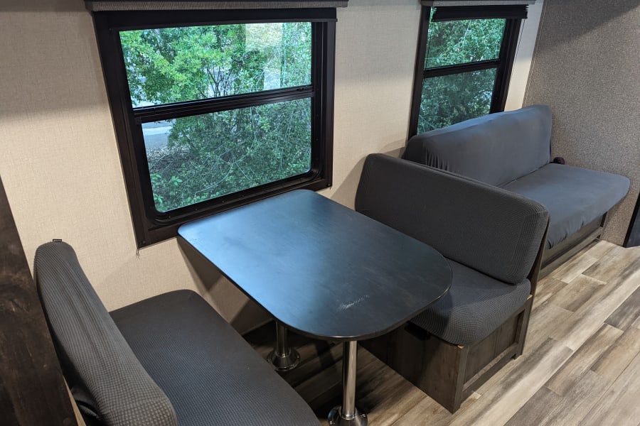 RV rental interior: dinette with dark table and benches, sofa, and windows with lush green views.