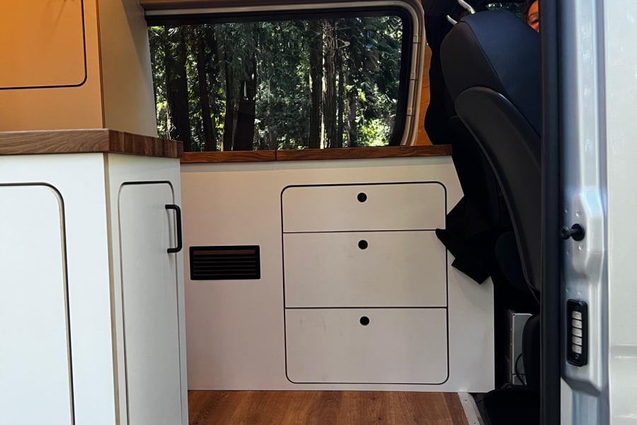 Modern RV rental interior: Clean white cabinets, wood paneling, ambient lighting, and a window view of trees.