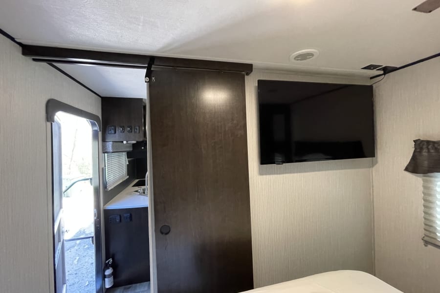 Inside a modern RV rental: a compact bedroom with a bed, a sliding door to the kitchen/galley, and a wall-mounted TV.