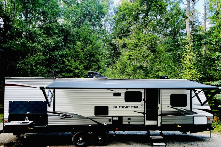 Discover this Pioneer travel trailer RV rental, featuring an extended awning & outdoor kitchen, nestled in a vibrant green forest.