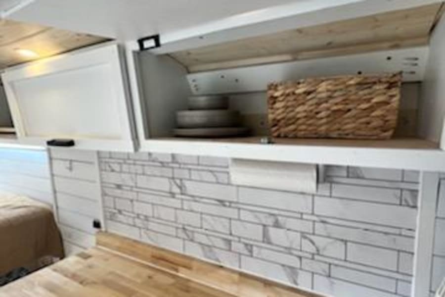 RV rental kitchen: Wood countertop, tile backsplash, & accessible overhead storage with dishes & a basket.
