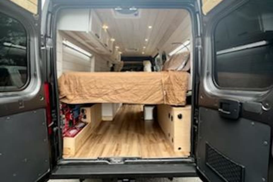 RV rental: Inside a converted van, a cozy bed, wooden floor, and bright paneled walls are visible through the open rear doors.