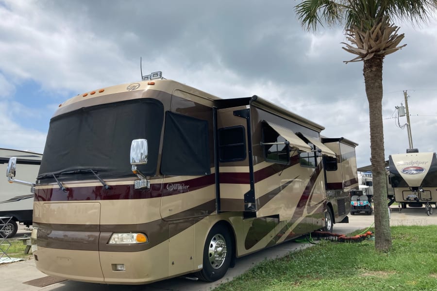 A large, two-tone tan and maroon RV rental motorhome, a Monaco Windsor, is parked at a campsite with awnings extended.