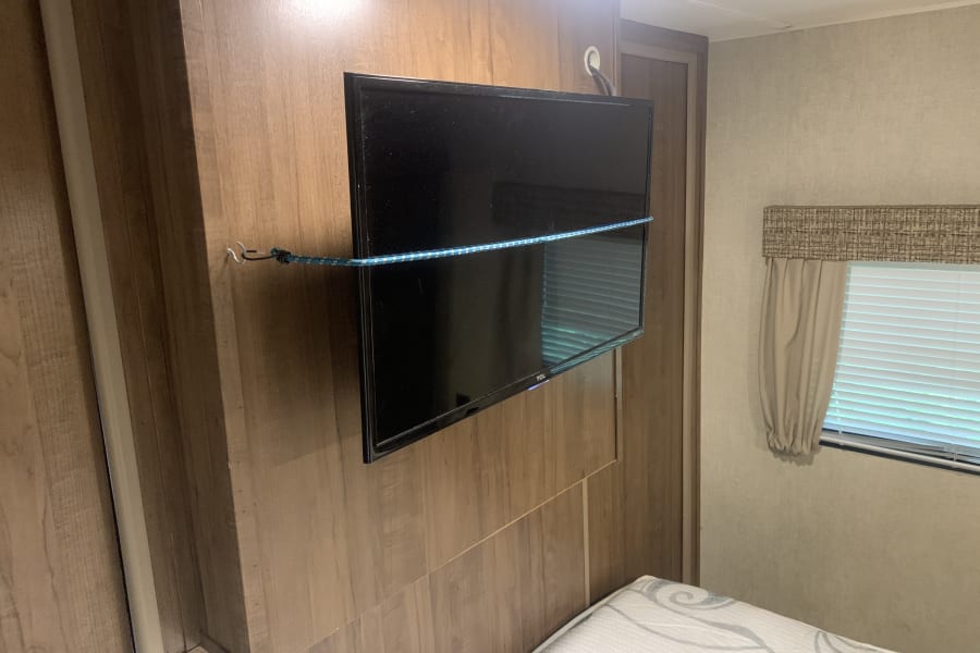 RV rental interior with a wall-mounted TV on wood paneling, secured by a blue bungee cord for travel. A bed and window are visible.