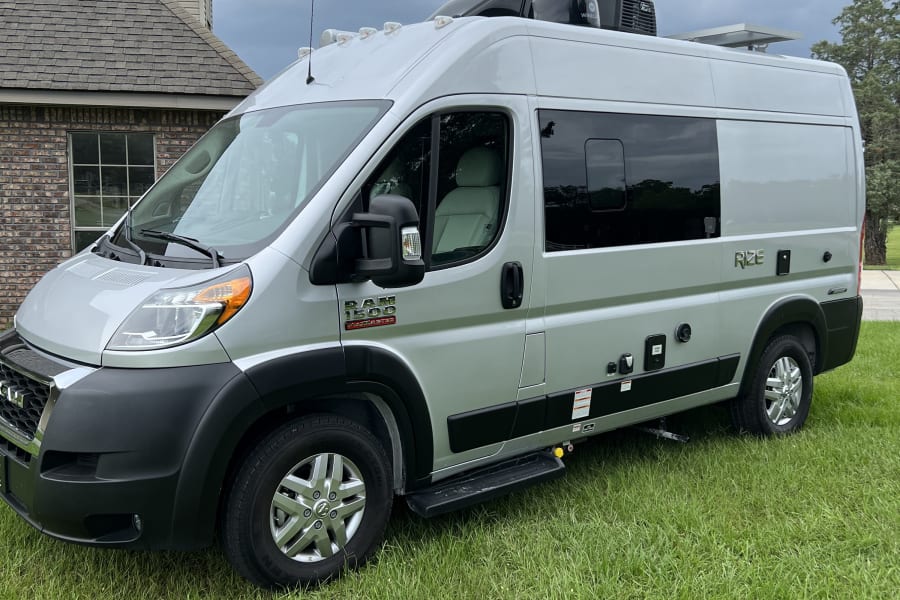Silver Ram ProMaster camper van parked on green grass, ready as an RV rental. Visible features include a deployed step and roof units.