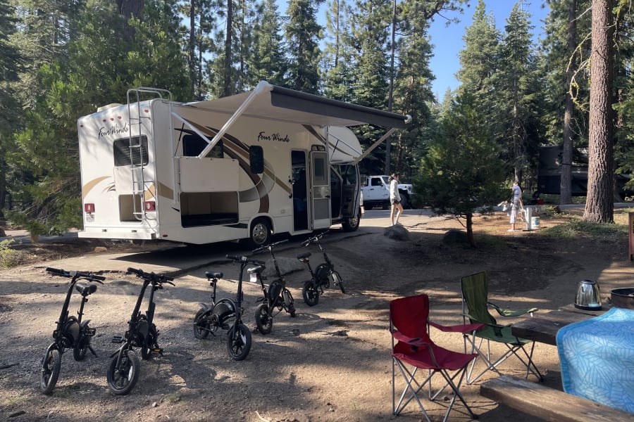 RV rental adventure: a "Four Winds" motorhome with awning extended, surrounded by trees at a campsite, with bikes parked.