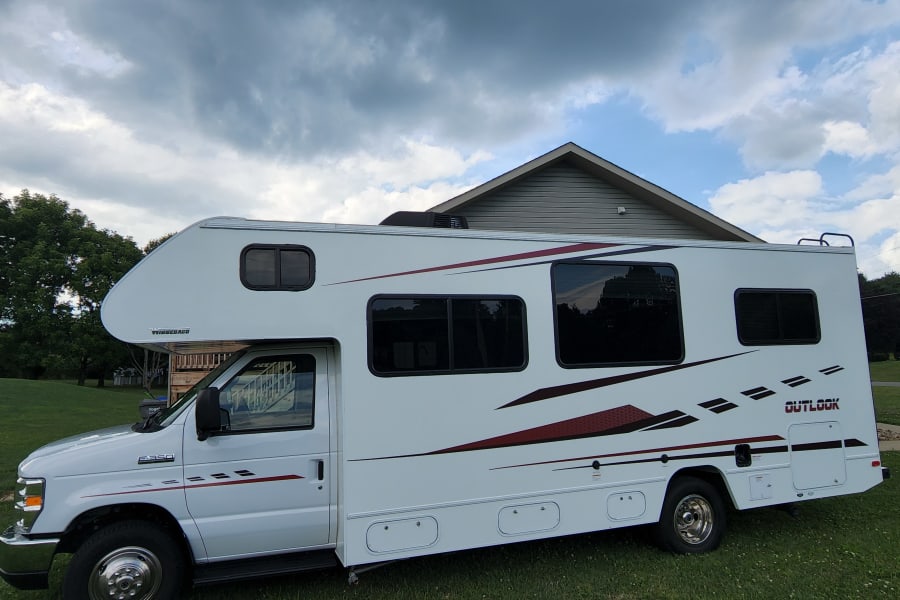 White Winnebago Outlook RV rental parked on grass by a house under a cloudy sky.