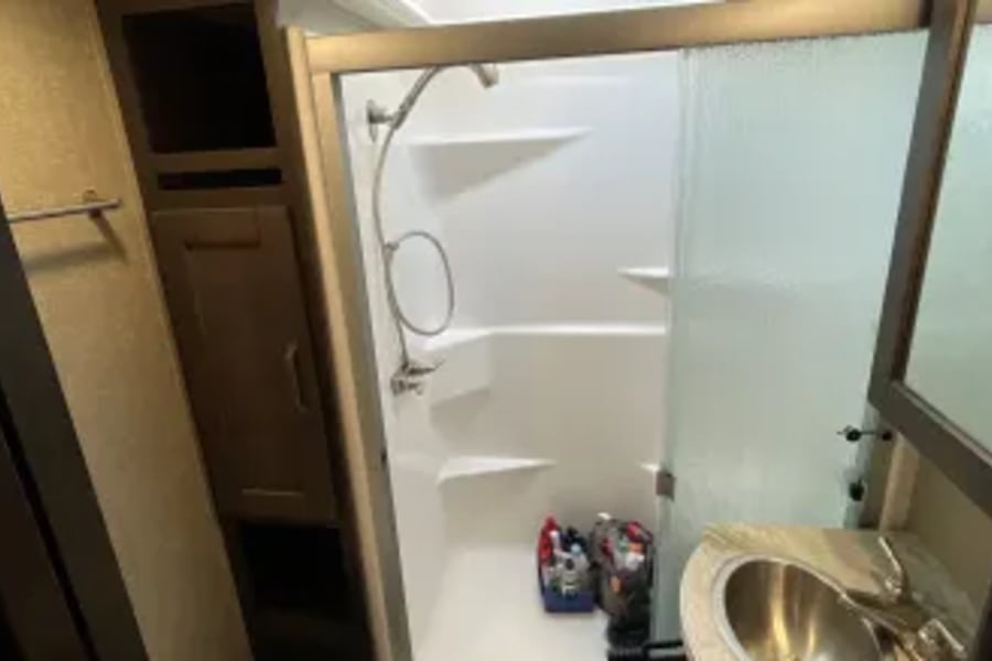 Inside an RV rental: a compact bathroom featuring a white shower with glass door, small sink, and wood storage, ideal for travel.