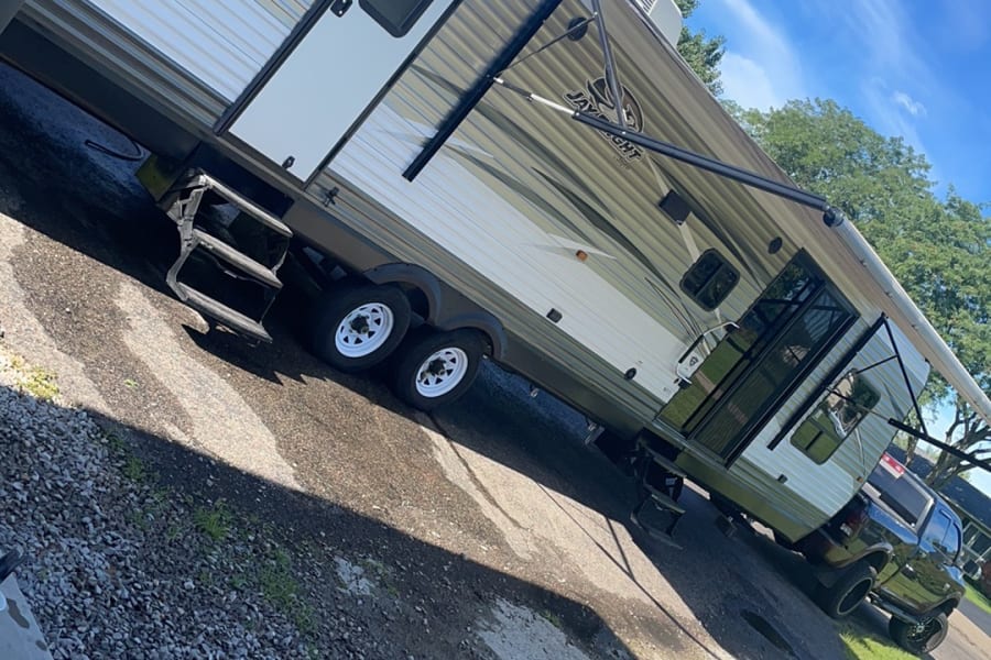 Bright travel trailer RV rental with awning extended, parked on gravel and concrete under a blue sky.