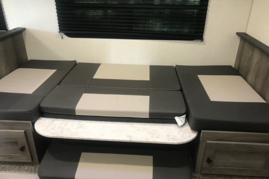 This RV rental features a dinette that converts into a bed, with grey and beige cushions, a table, and built-in storage.