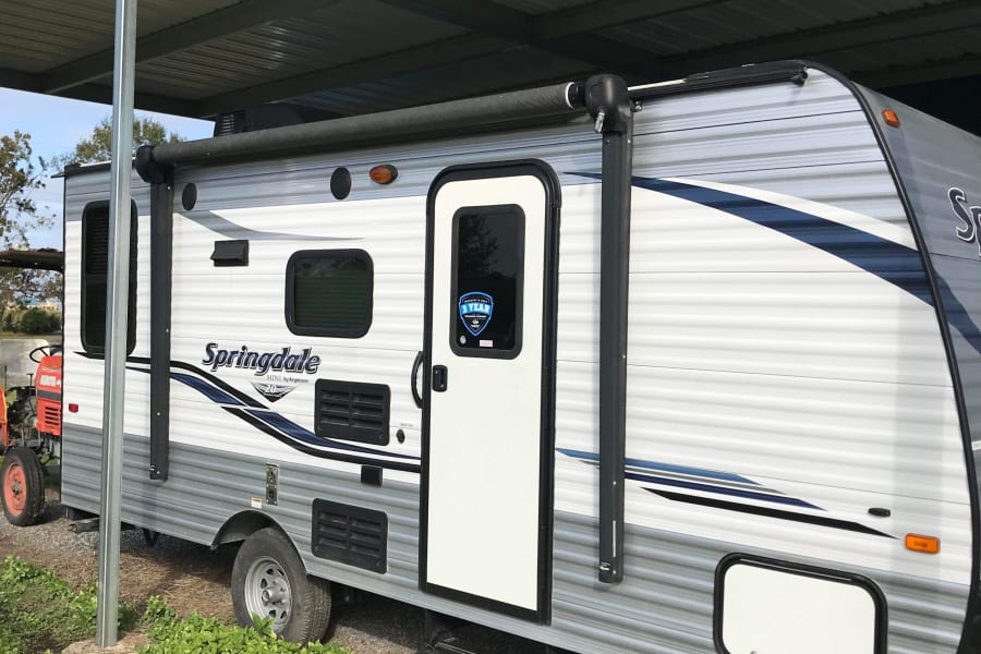 A white and gray Springdale travel trailer, a potential RV rental, is parked under a metal carport with its awning extended.