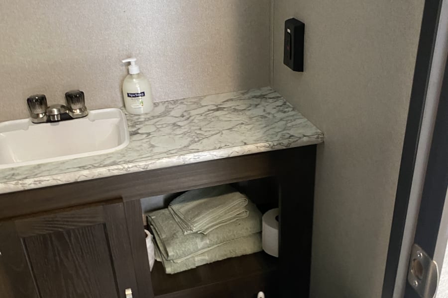 Inside an RV rental bathroom: compact sink, marble counter, dark vanity with stored towels & toilet paper, and a toilet.