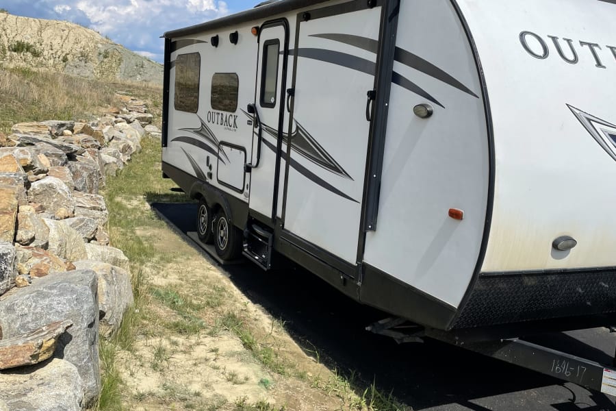 White Outback travel trailer, perfect for an RV rental, parked beside a rock wall and house on a bright, sunny day.