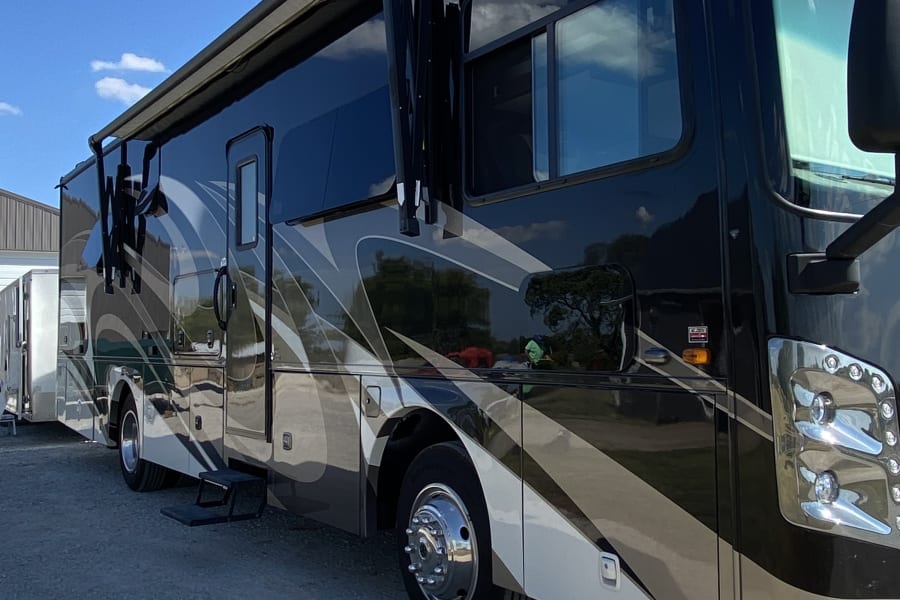 Stunning black and tan motorhome for rv rental, parked on gravel with an awning extended, under a bright blue sky.