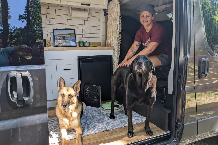 A happy woman pets her black Lab, while a German Shepherd rests in their spacious rv rental, with the door open to nature.