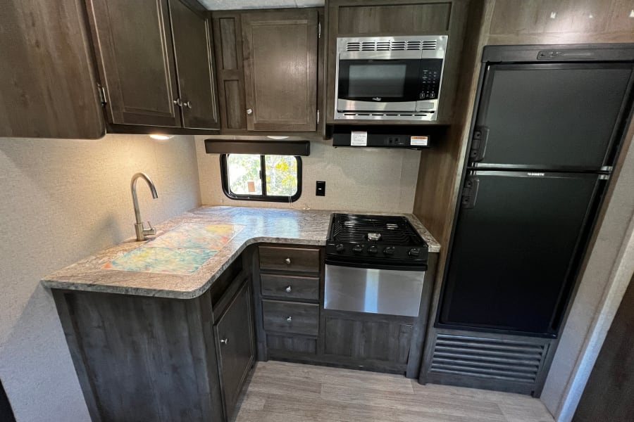 Interior of a functional RV rental kitchen with dark wood cabinets, microwave, stove, refrigerator, and sink.