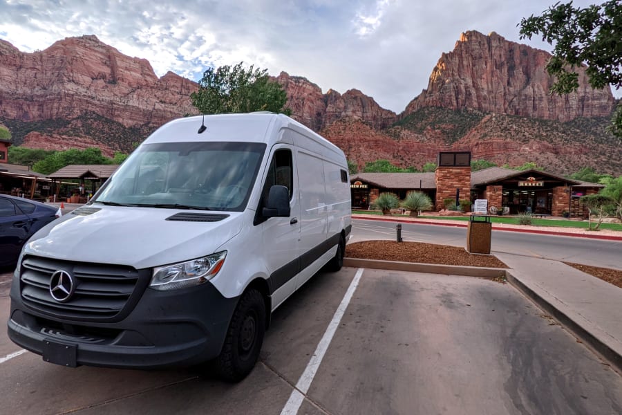 White Mercedes Sprinter RV rental parked in Zion National Park, with towering red mountains and village buildings in the background.