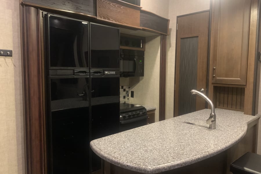 Well-equipped RV rental kitchen: large black fridge, microwave, stove, and a granite-look curved counter with sink.