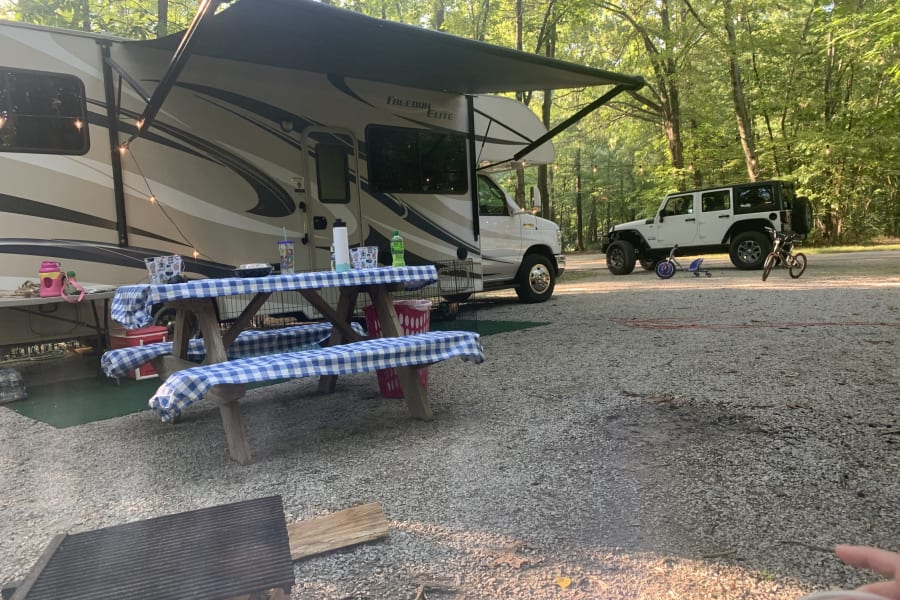 Enjoying a family campsite! This RV rental set up with a picnic and grill under string lights, plus a Jeep and bikes, offers outdoor fun.