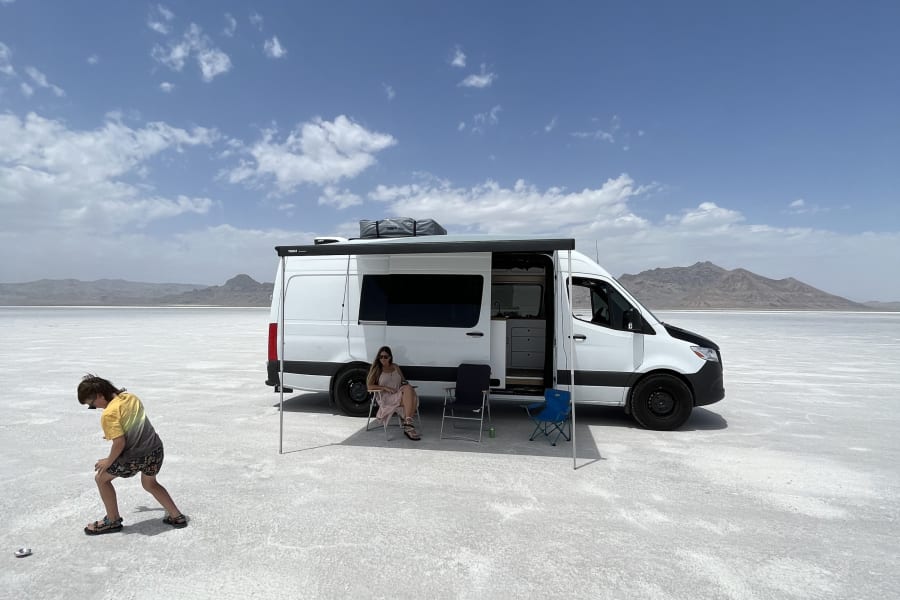 RV rental on vast salt flats under a blue sky. A woman relaxes under the awning, a child plays nearby. Road trip fun!
