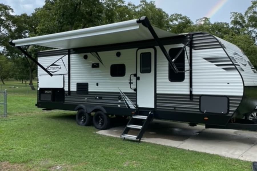 Experience camping with an RV rental! This white travel trailer with an extended awning is parked by grass under a double rainbow sky.