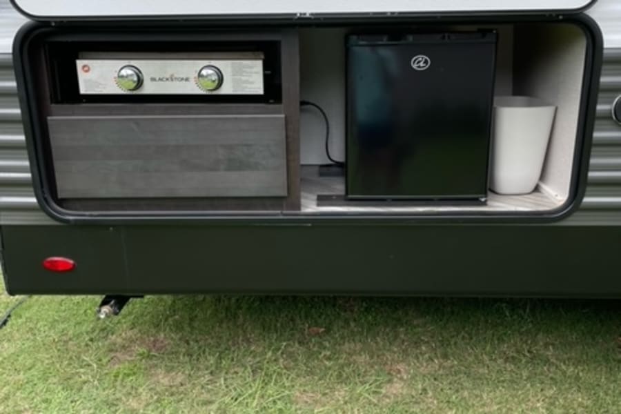 RV rental showing its outdoor kitchen: a mini-fridge, Blackstone griddle, and counter built into the side, on grass.