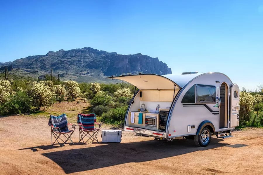 RV rental ready for adventure: Teardrop trailer with open kitchen, chairs, & desert mountains.