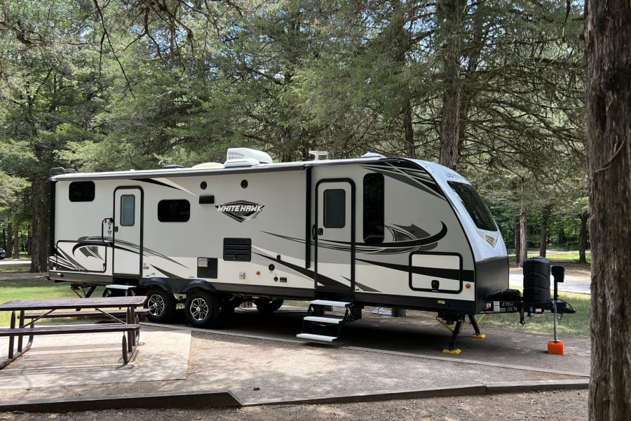 RV rental: A light grey and black White Hawk travel trailer is parked at a serene, tree-lined campsite with a picnic table.