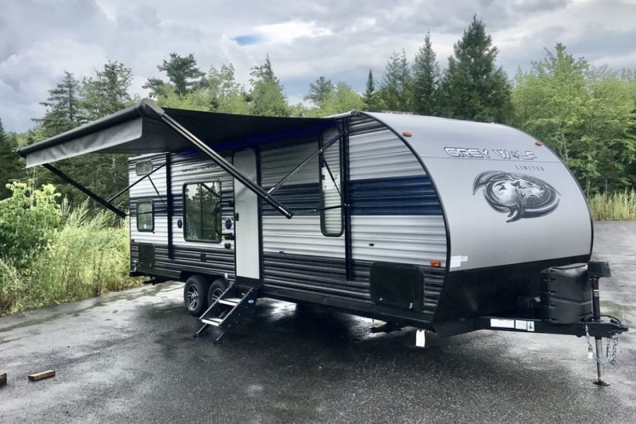 Grey travel trailer RV rental with awning extended, parked on a wet lot by trees under a cloudy sky. Perfect for your trip.