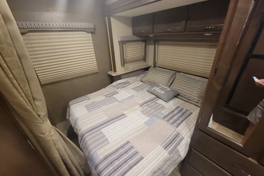 RV rental bedroom: Cozy interior with bed, window blinds, patterned valances, and overhead cabinets.